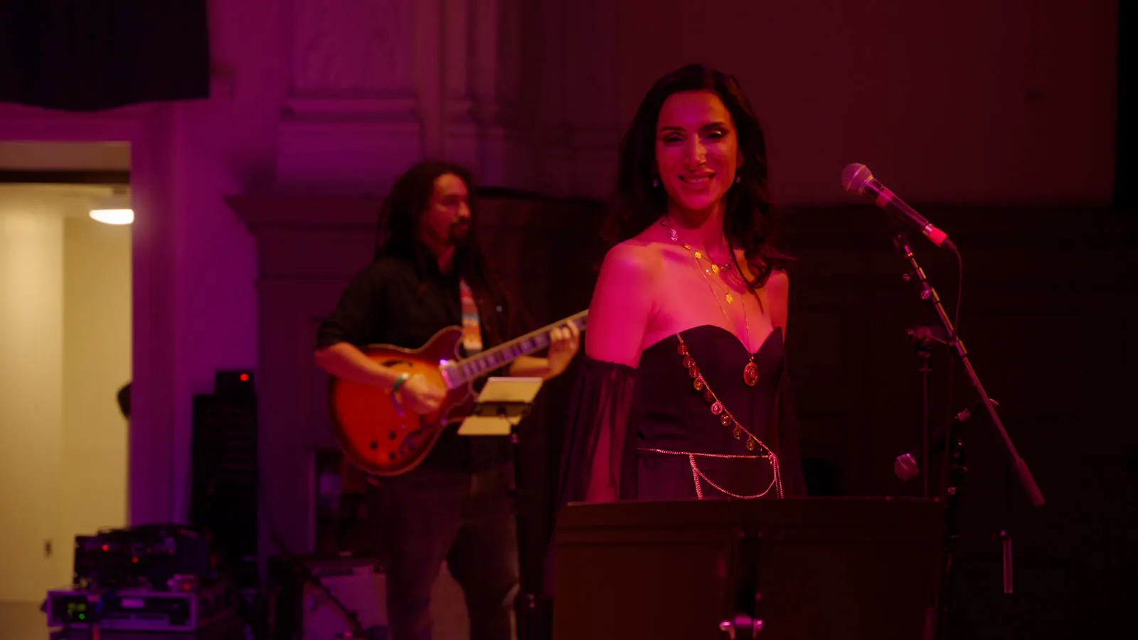 Sura smiling at the microphone in magenta stage light, guitarist behind her