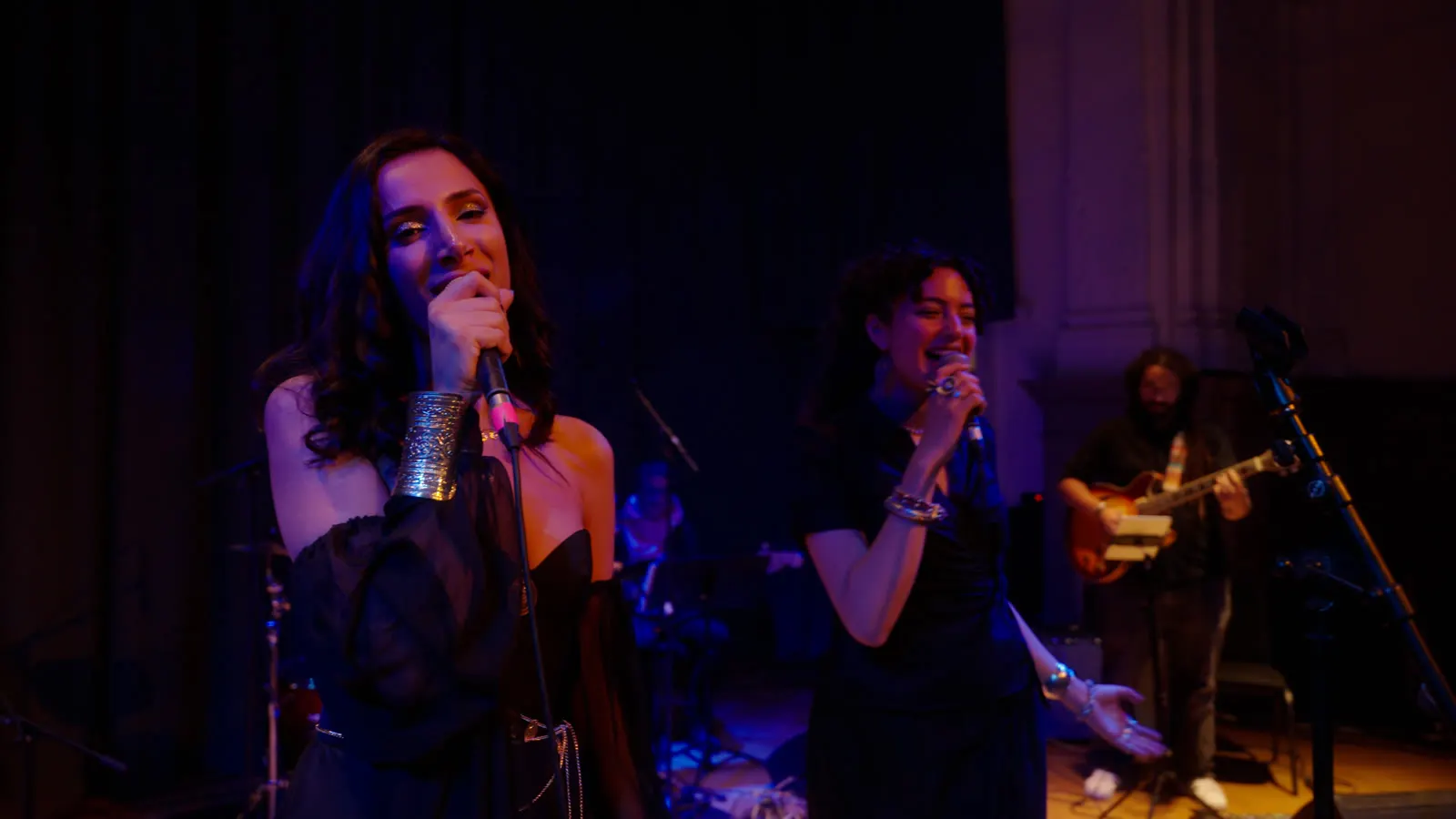 Sura singing a duet with a second vocalist in blue-violet light